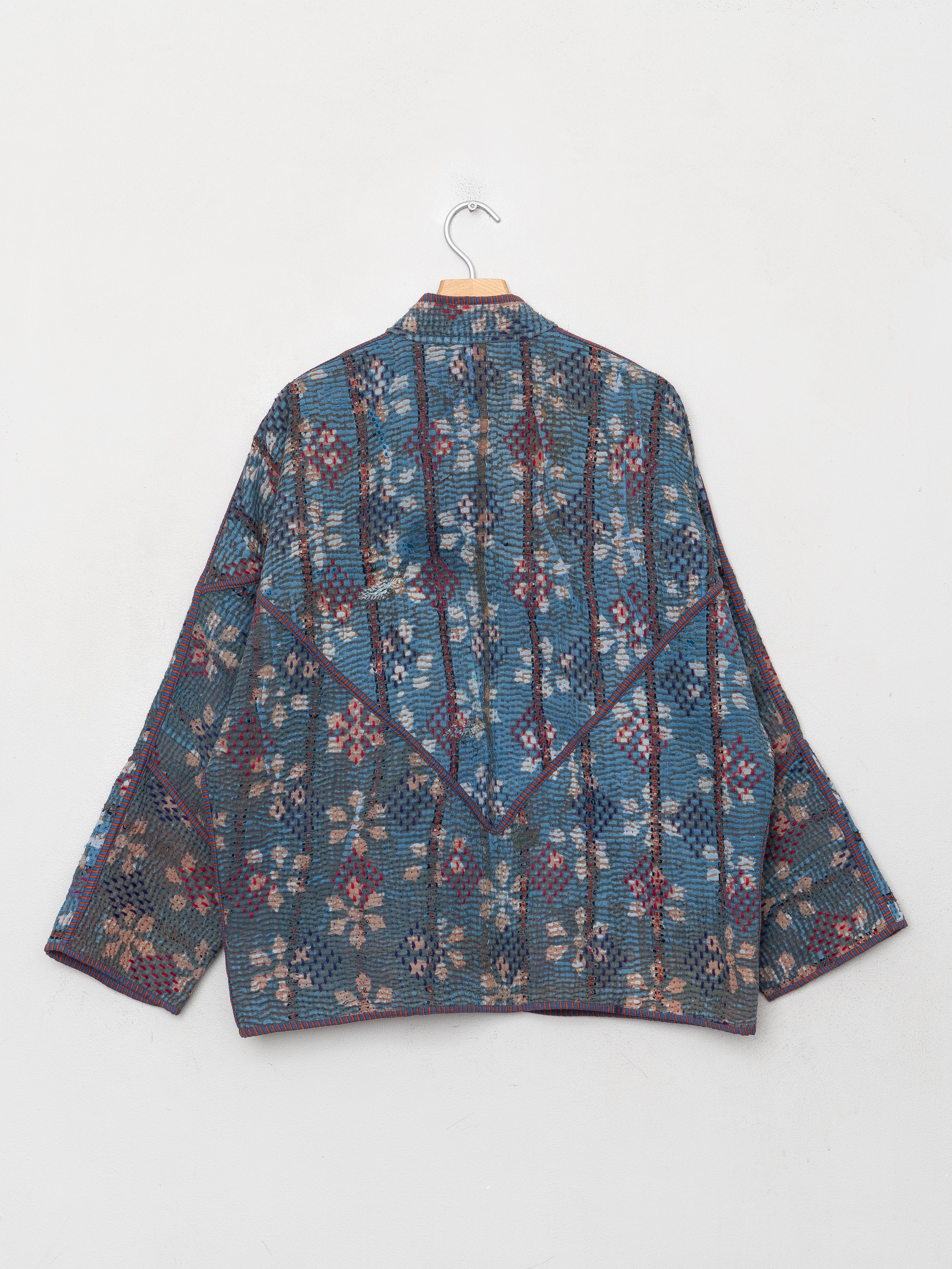 A long-sleeve patterned jacket with a blue, red, and beige geometric and floral design, hanging on a wooden hanger against a plain light gray wall.